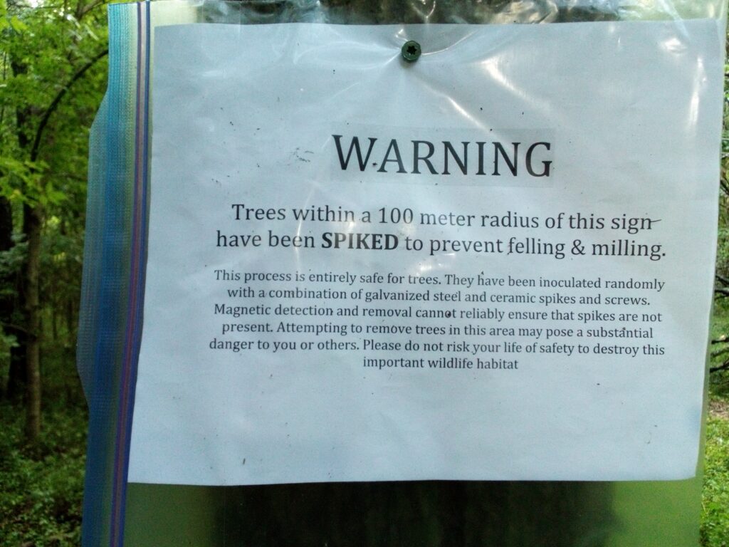 sign affixed to a tree that reads "WARNING Trees within a 100 meter radius of this sign have been SPIKED to prevent felling & milling. This process is entirely safe for trees. They have been inoculated randomly with a combination of galvanized steel and ceramic spikes and screws. Magnetic detection and removal cannot reliably ensure that spikes are not present. Attempting to remove trees in this area may pose a substantial danger to you or others. Please do not risk your life or safety to destroy this important wildlife habitat."