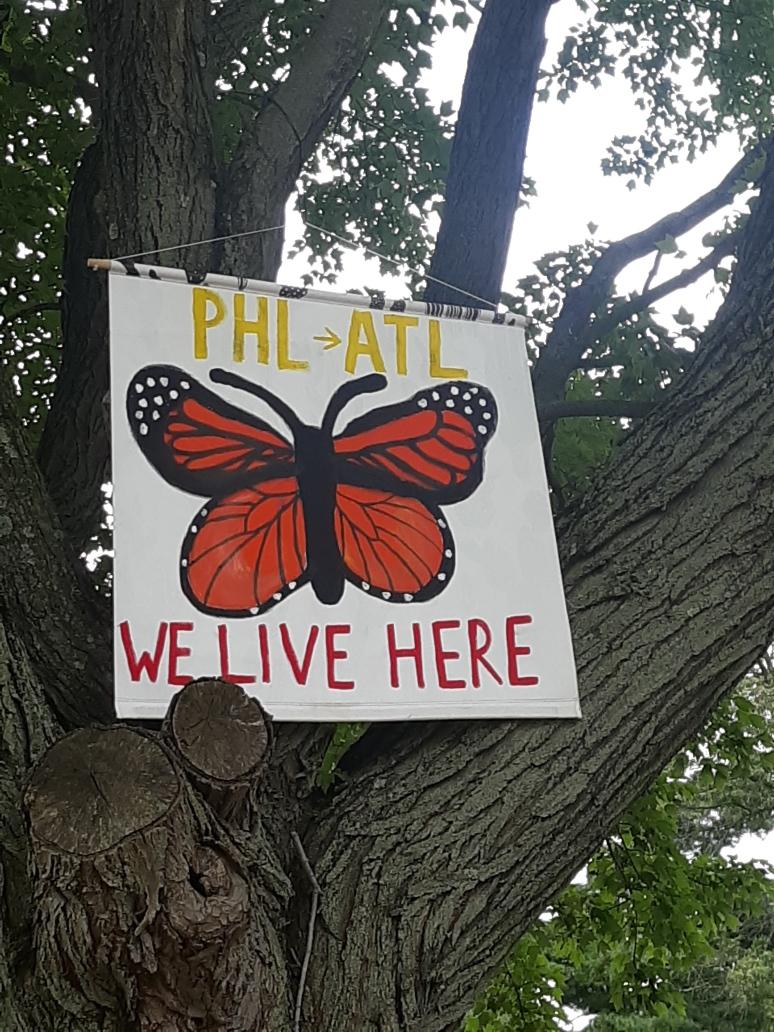 A close-up picture of a banner in a tree reads "PHL to ATL, WE LIVE HERE" with a monarch butterfly in the middle. 