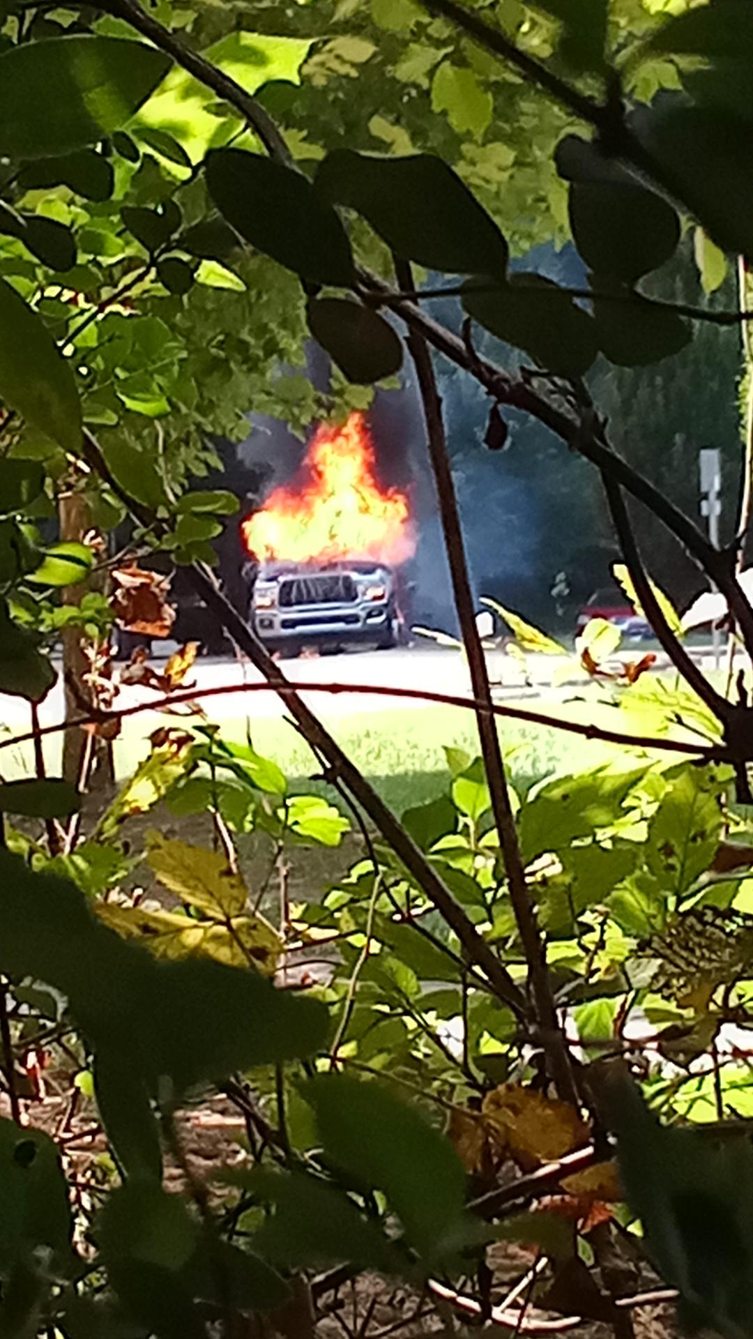 Through the leaves of a tree, a truck is seen burning in a gravel parkng lot.
