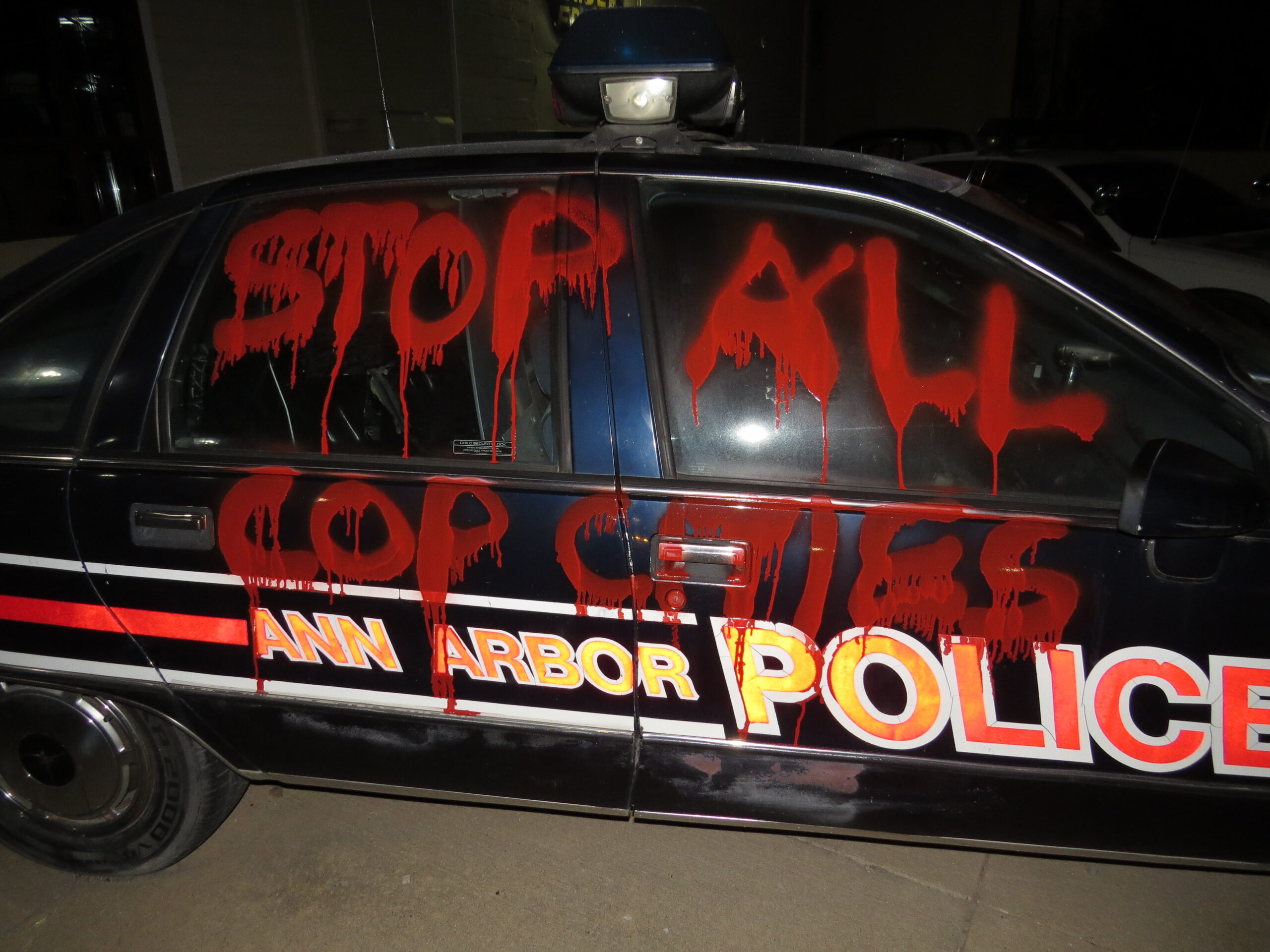 An Ann Arbor police car is spray painted "STOP ALL COP CITIES" on the side in red spray paint.