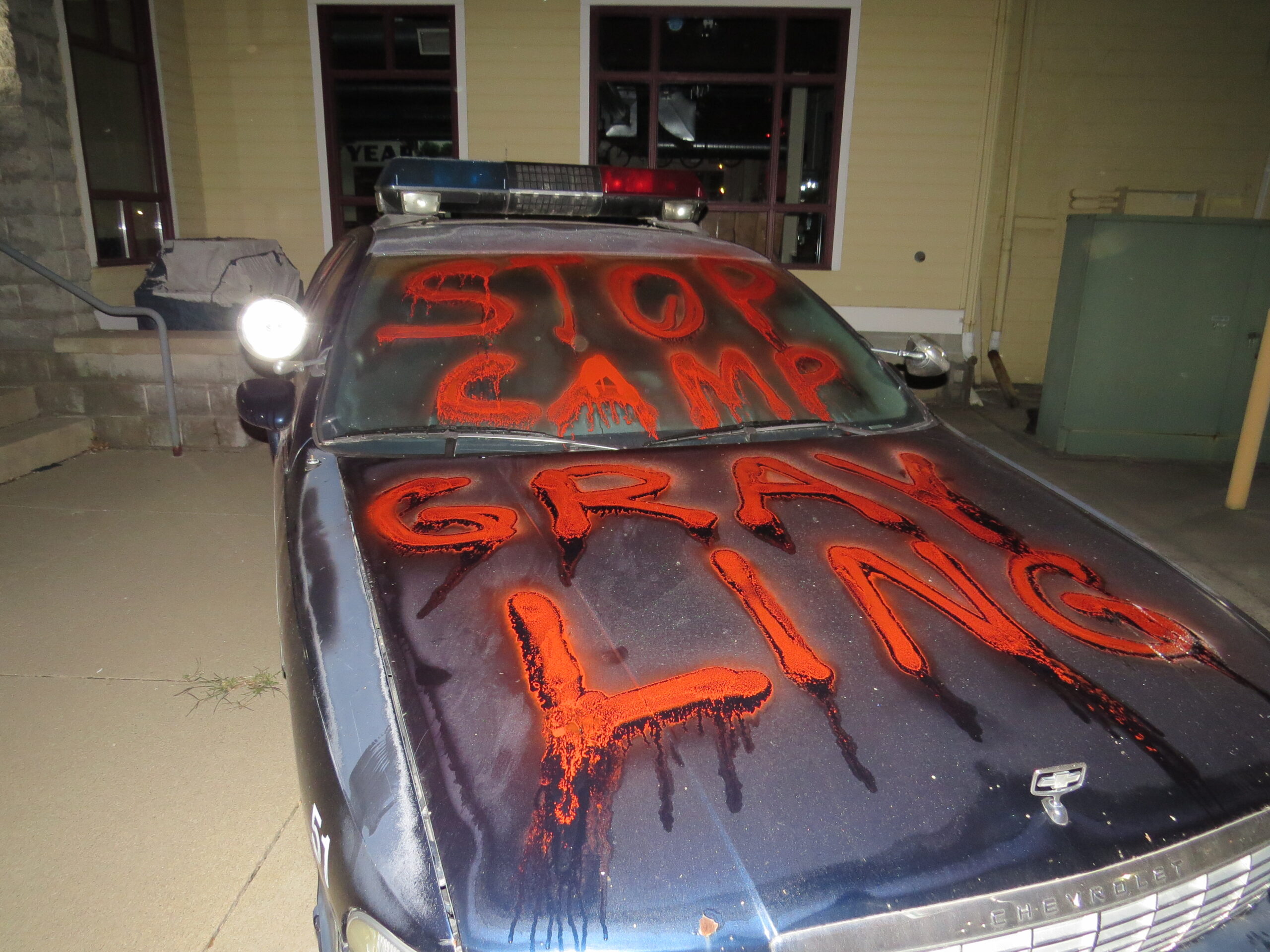 A police car is spray painted "STOP CAMP GRAYLING" in red spray paint.