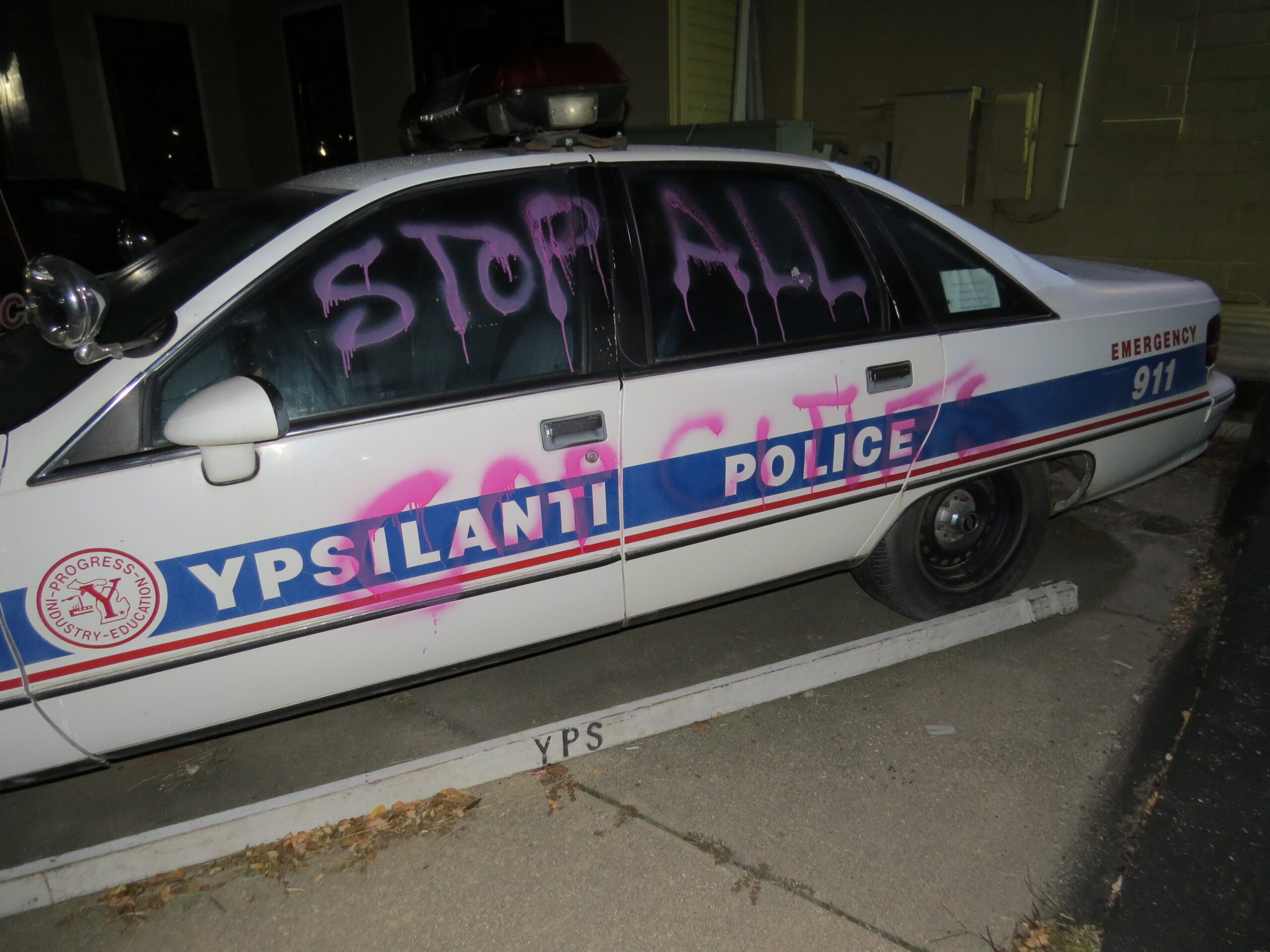 A Ypsilanti police car is spray painted "STOP ALL COP CITIES" on the side in pink spray paint.