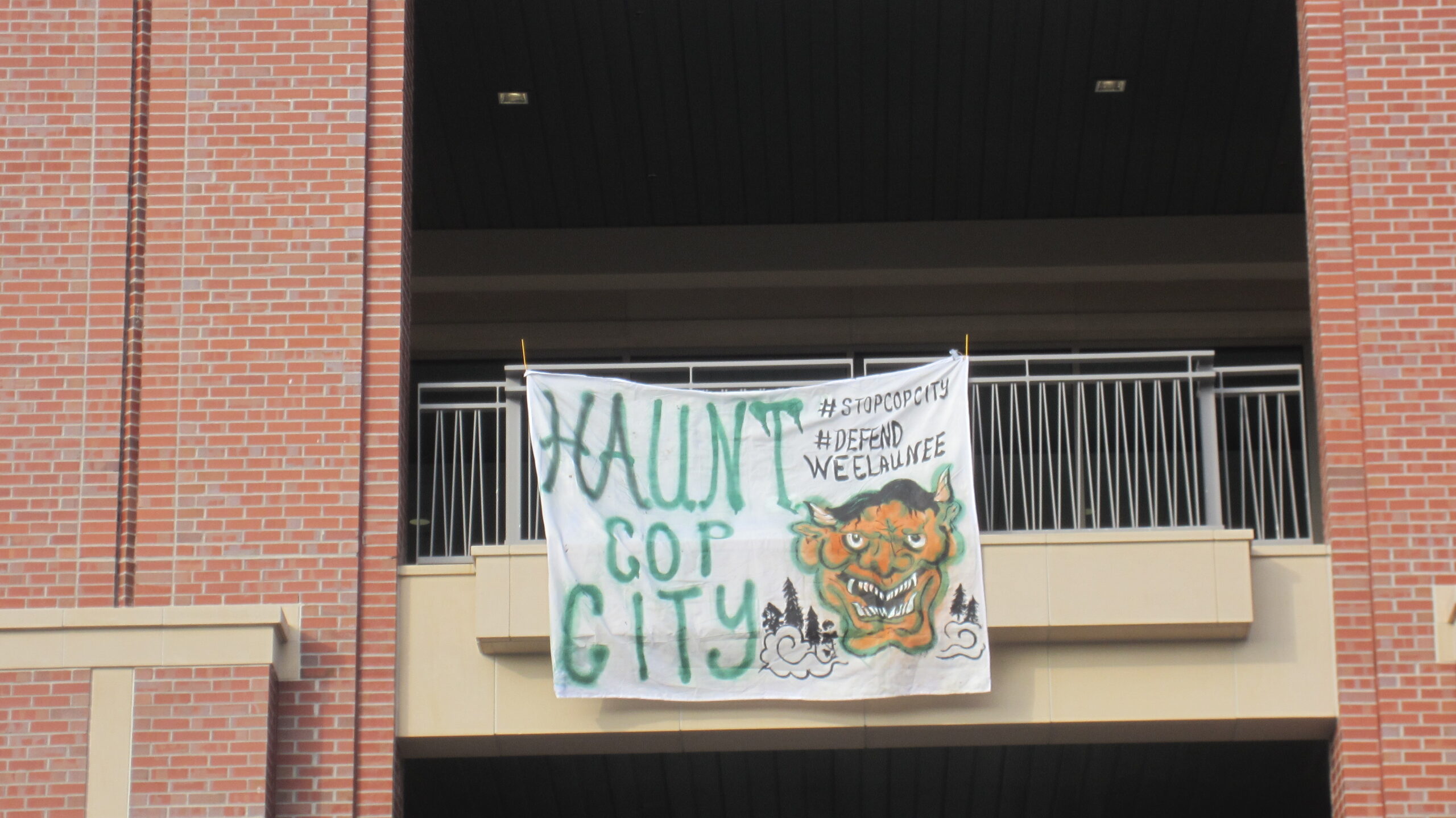 A banner hung on a railing reads "HAUNT COP CITY" in big green letters and "#stopcopcity, #defendweelaunee in smaller black letters. A Hannya mask face with trees and wind are depicted below it.