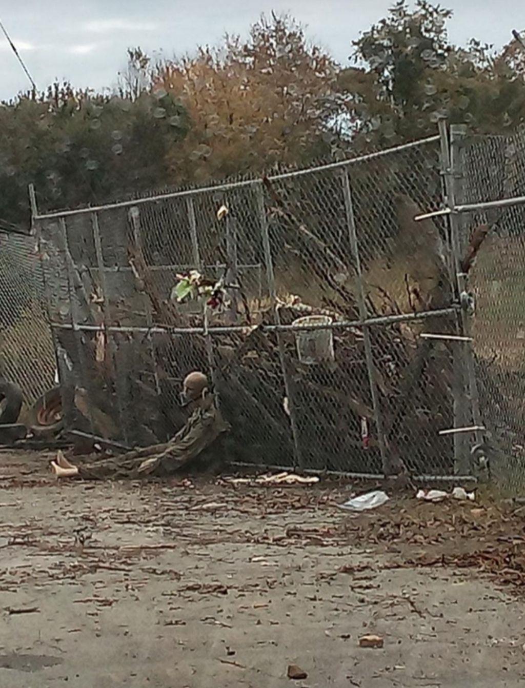 A mannequin dressed in green military-esque coveralls sits propped up against a fence reinforced with branches and old car tires. The mannequin's neck head is partially severed, and there is a U-Lock around its neck.