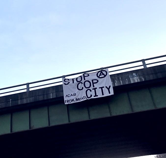 A white banner with black text hangs on a bridge that reads "STOP COP CITY ACAB FROM AVL to ATL" with an anarchist circle-A symbol.