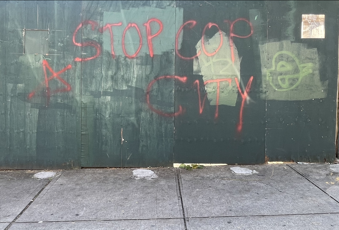 "STOP COP CITY" and a stylized "A" without a circle around it spray painted in red paint on a green wall in front of a sidewalk