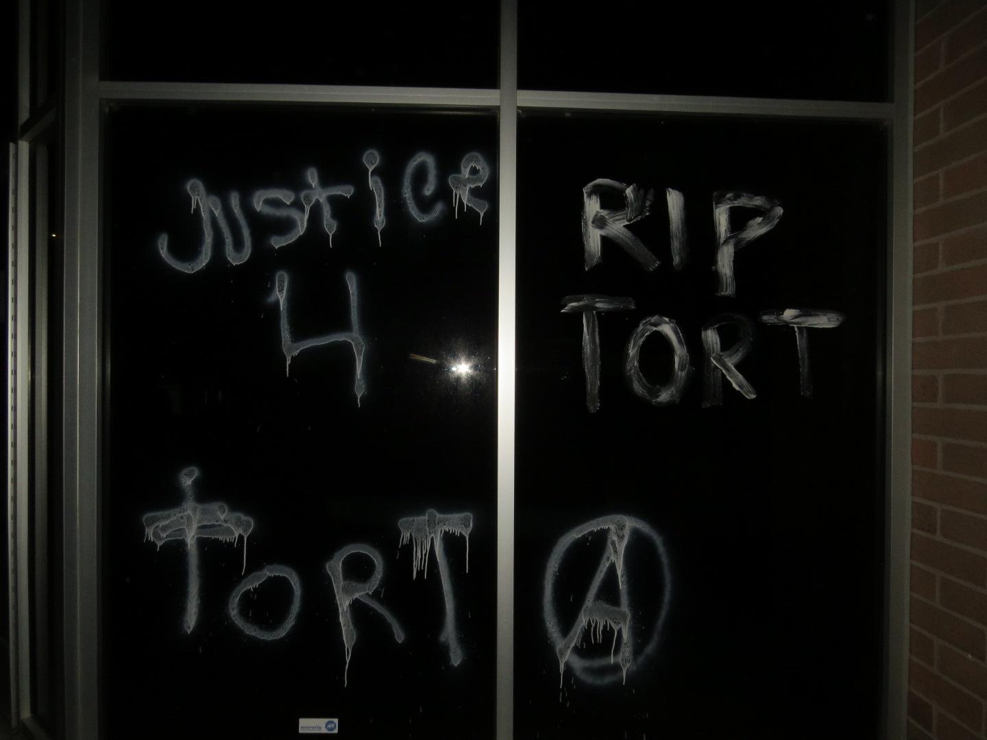 On a black window, white paint spells "JUSTICE 4 TORT" on one pane, and "RIP TORT" with an anarchist circle-A symbol on the other pane.