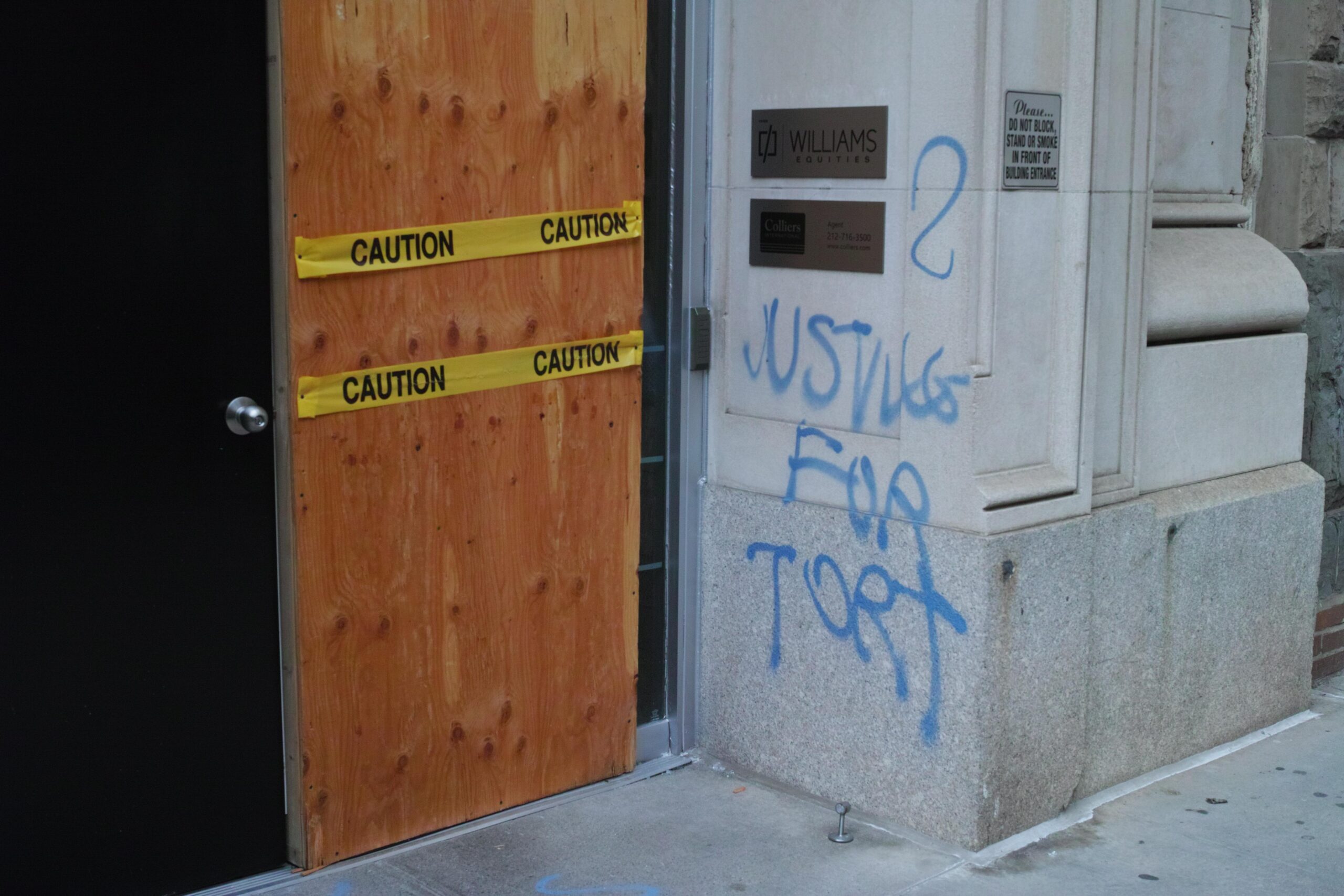 Onto a concrete wall, "JUSTICE FOR TORT" is tagged in blue spray paint. A door next to the wall is covered in plywood and caution tape.