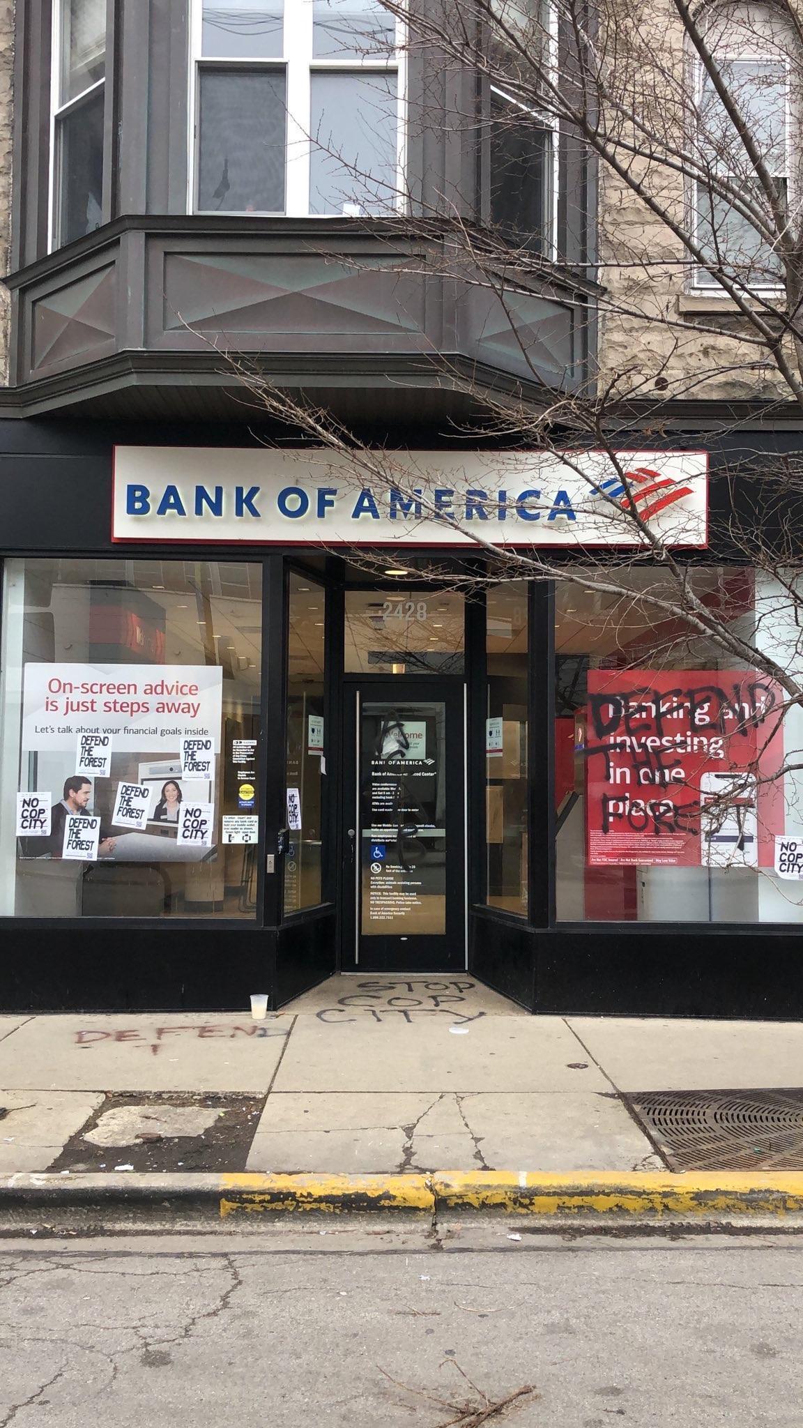 On a city street, a Bank of America bank has tags that say "Defend the Forest" and "Stop Cop City" in black spray paint.