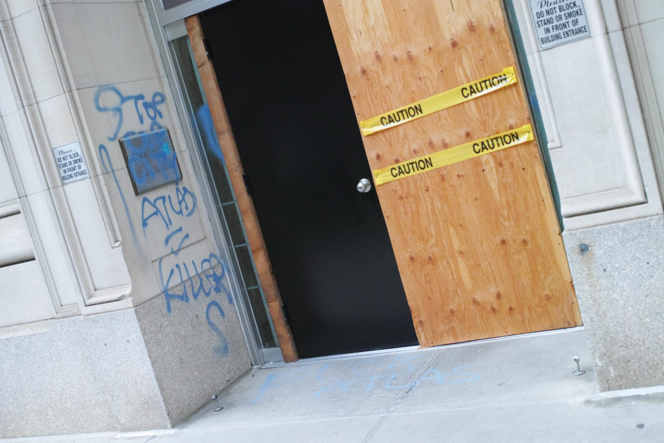 Onto a concrete wall, "STOP COP CITY ATLAS = KILLERS" is tagged in blue spray paint. On the sidewalk, "FUCK ATLAS" is also tagged in blue spray paint. A door next to the wall is covered in plywood and caution tape.