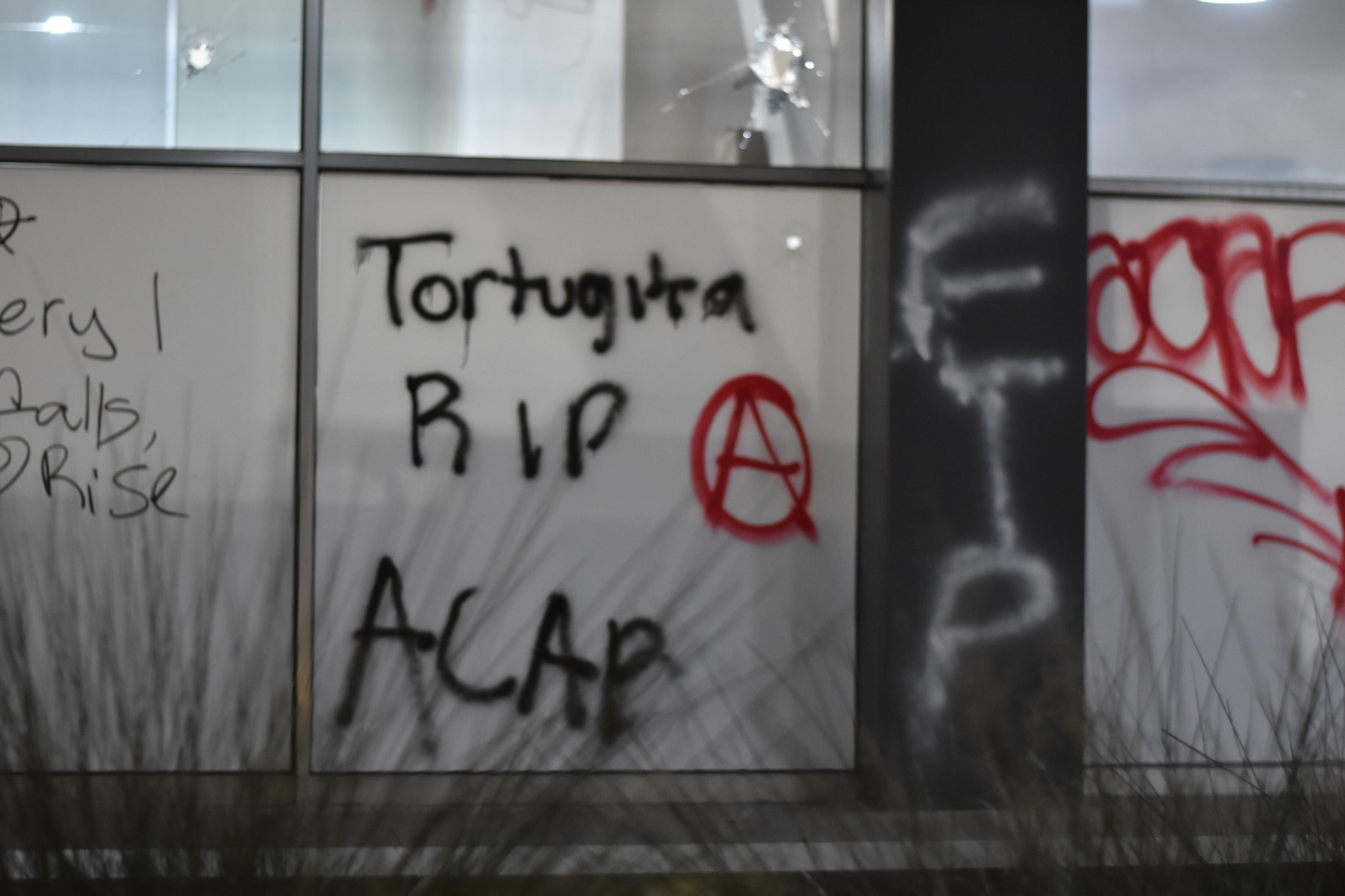 Onto a wall, "Tortuguita RIP" and "ACAB" are tagged in black spray paint. "FTP" is tagged in white spray paint. "ACAB" and an anarchist circle-A symbol are tagged in red spray paint.