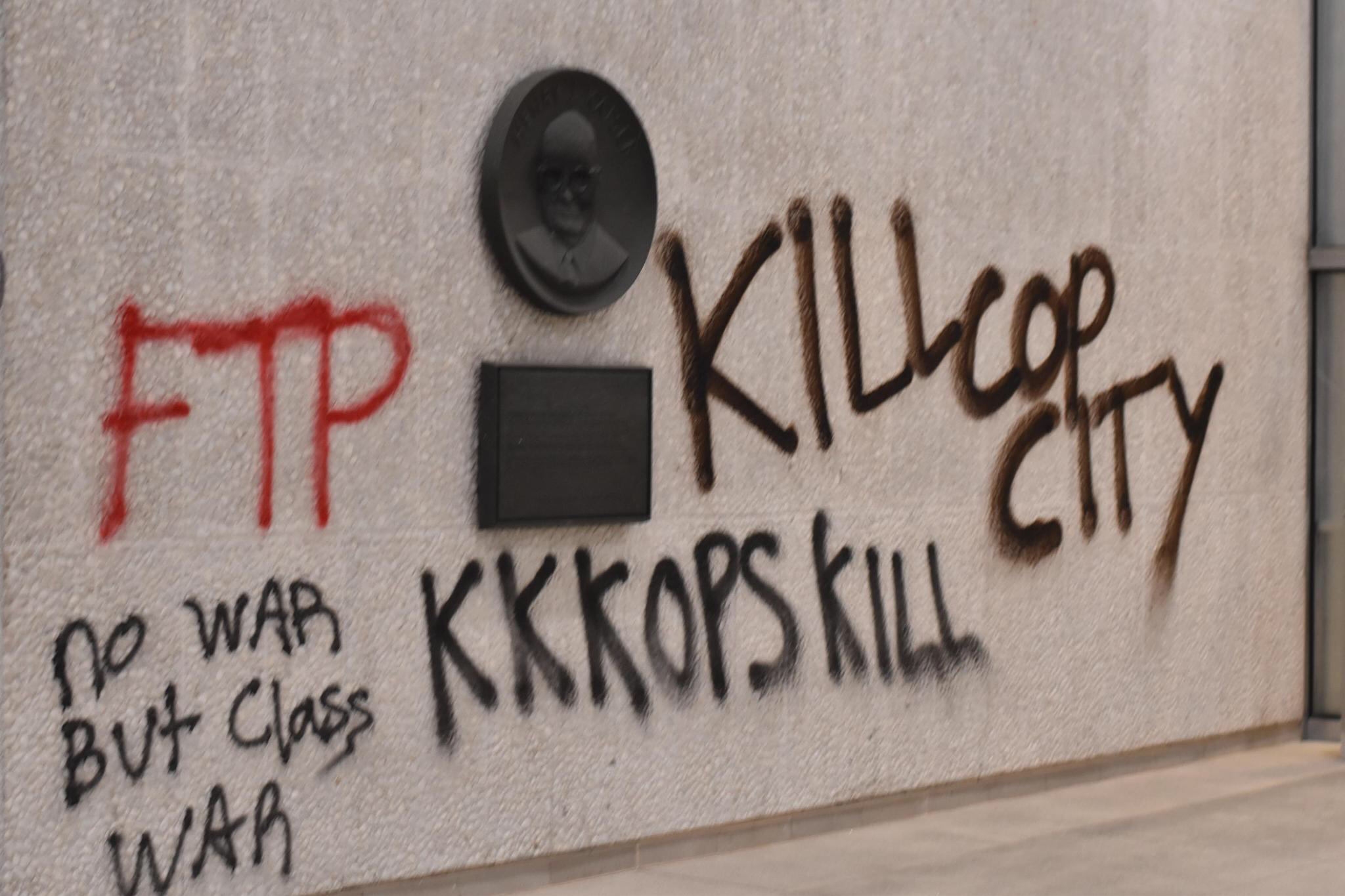 Onto a concrete wall, "FTP" is tagged in red spray paint. "no war but class WAR," "KKKOPS KILL," and "KILL COP CITY" is  tagged in black spray paint.