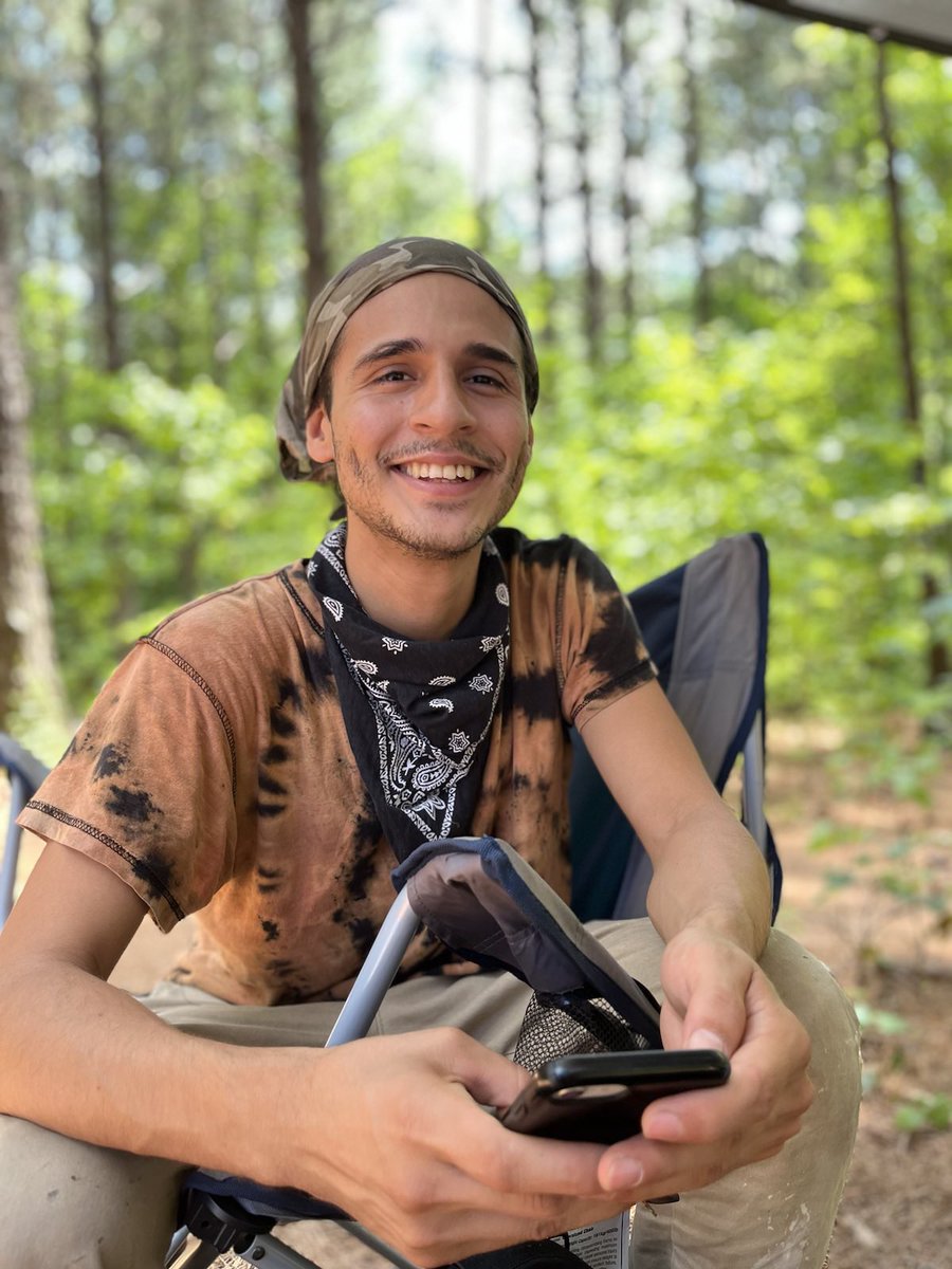 A forest defender wearing a tie die brown and black shirt and wearing a black bandana around its neck sits in a folding chair in a dense forest, smiling.