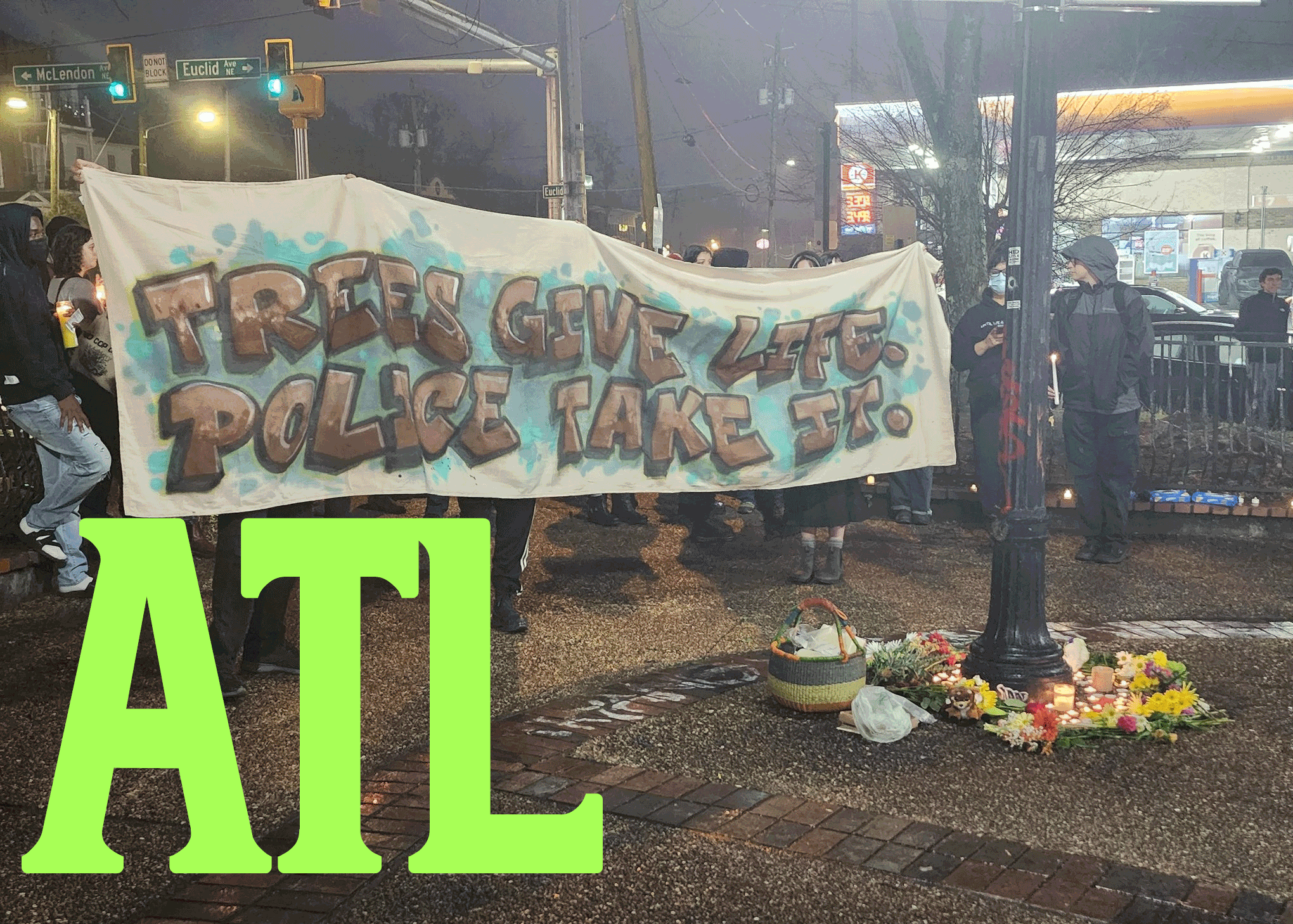 A banner that states "TREES GIVE LIFE POLICE TAKE IT." In purple letters with blue backsplash is held by several vigilers. On the left is a light pole surrounded with flowers and candles. In the bottom left corners, "ATL" is spelled in green letters.