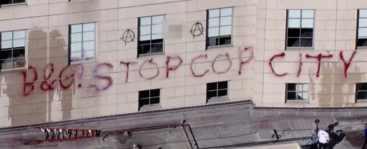 "B&G! STOP COP CITY" is sprayed on the side of a building with several windows in red spray paint. Several people standing nearby appear to be attempting to clean the paint with little success.