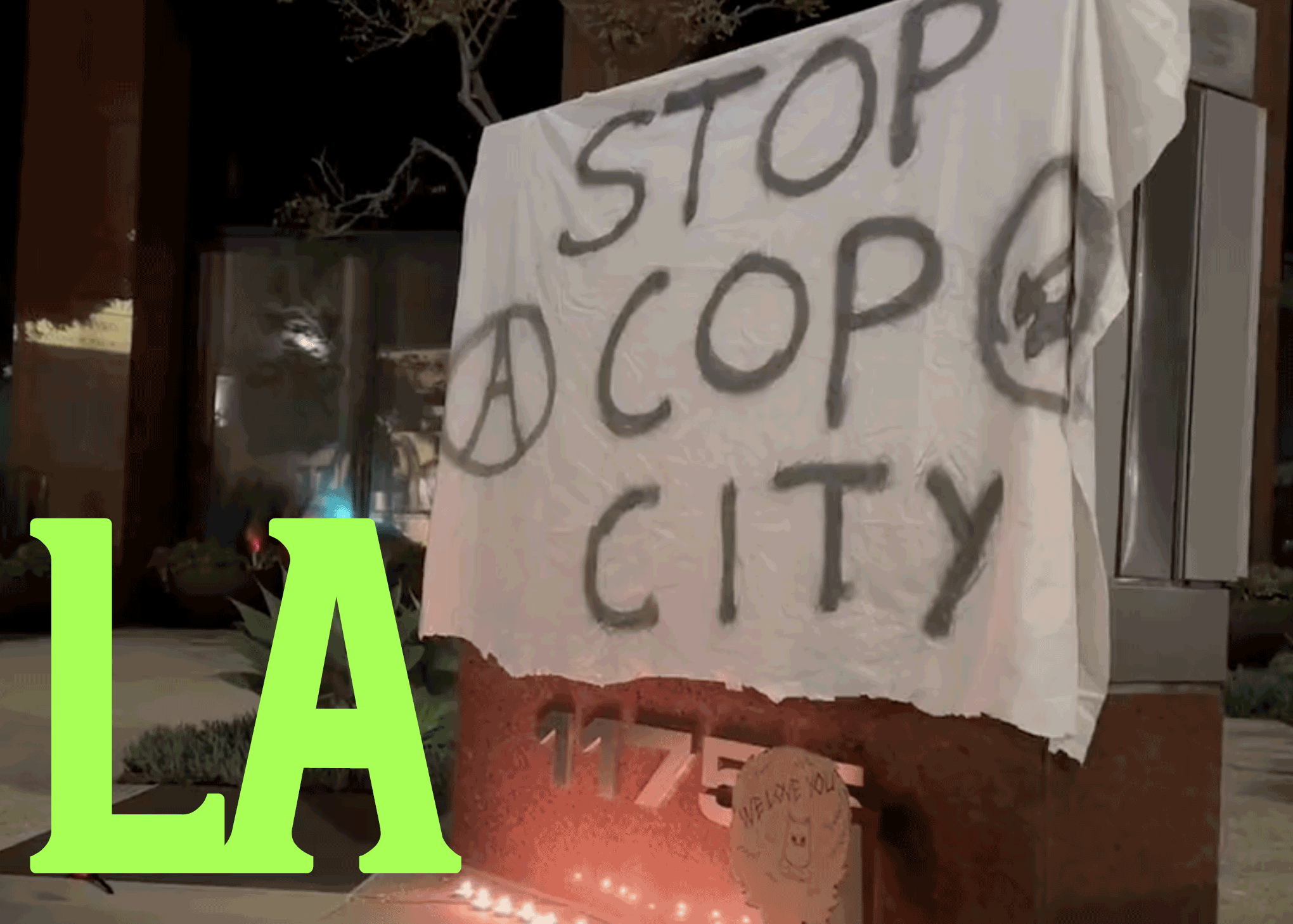 A white banner with black text spells out "STOP COP CITY" with an anarchist circle-A and a three arrows symbol in a circle. In the bottom left corners, "LA" is spelled in green letters.