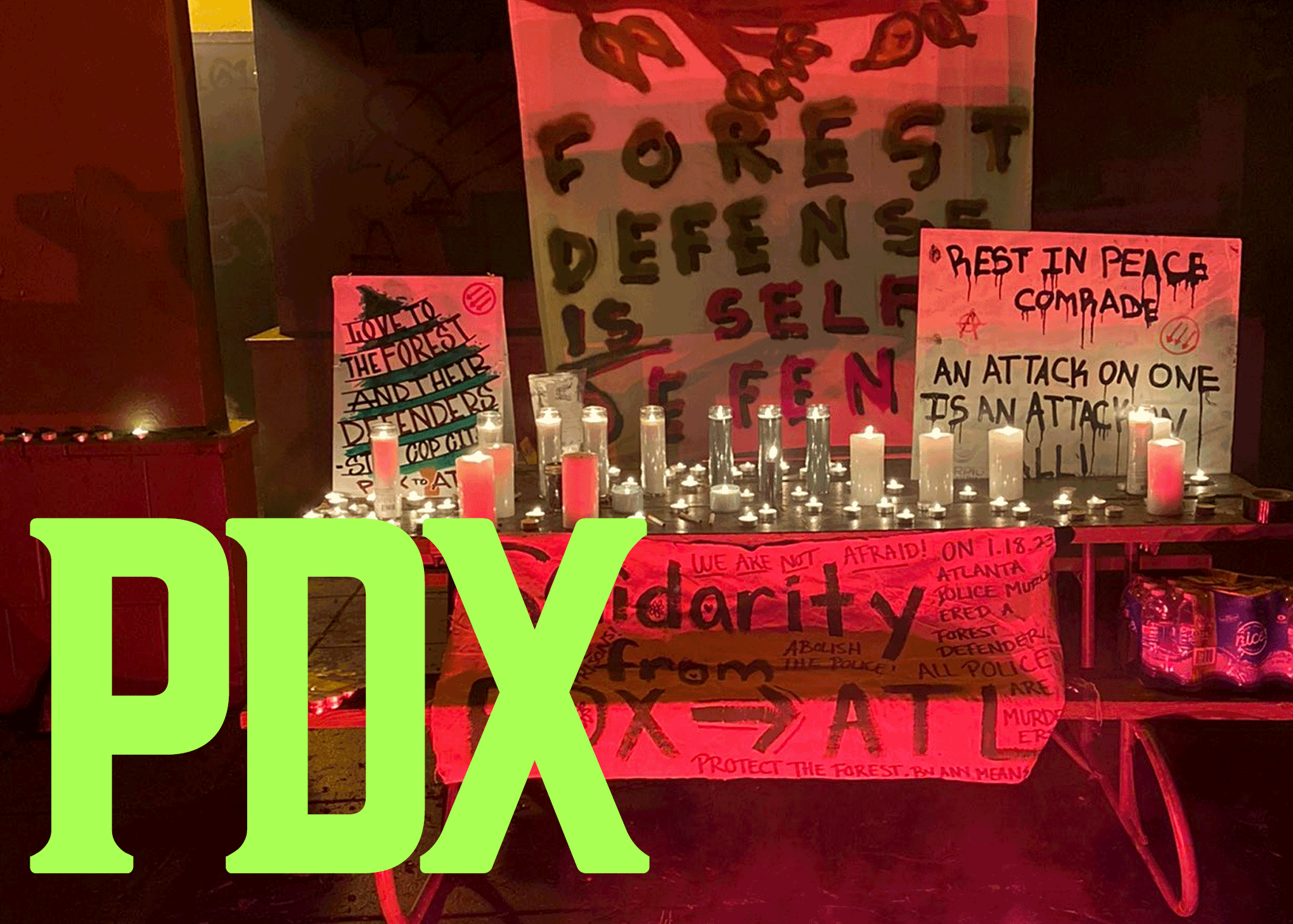Three signs and a banner are lit in red on a picnic table filled with candles. The first sign is styled as a tree and states "LOVE TO THE FOREST AND THEIR DEFENDERS PDX TO ATL. The sign has a red three-arrows symbol in a circle. The second sign says "FOREST DEFENSE IS SELF DEFENSE" in green and red letters. The top of the sign is a tree branch. The third sign says "Rest in peace comrade, an attack on one is an attack on all!" A red anarchist circle-A symbol and a red three-arrows symbol adorn the sides of the sign. The banner is laid on the seat of the picnic table and has numerous messages on it. The largest states "Solidarity from PDX (arrow) ATL. The banner also states the following messages in smaller text in the margins of the larger message: "We are not afraid!" "Abolish the police!" "Protect the forest by any means." "All police are murderers." "On 1.18.23 Atlanta Police murdered a forest defender!" In the bottom left corners, "PDX" is spelled in green letters.