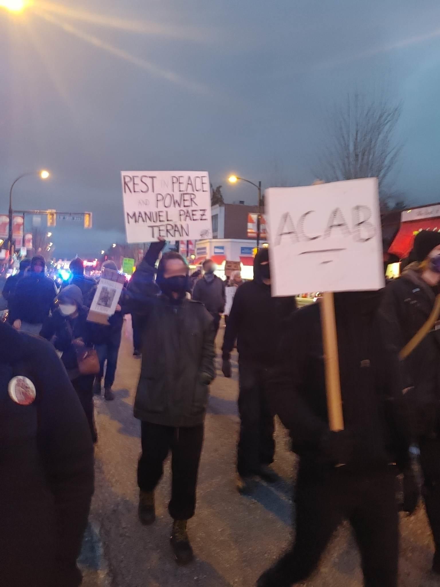 A group of protesters holding signs march on a city street. One sign says "REST IN POWER MANUEL PAEZ TERAN" and another says "ACAB", and is underlined twice. The faces of the protesters are blurred. It is twilight.