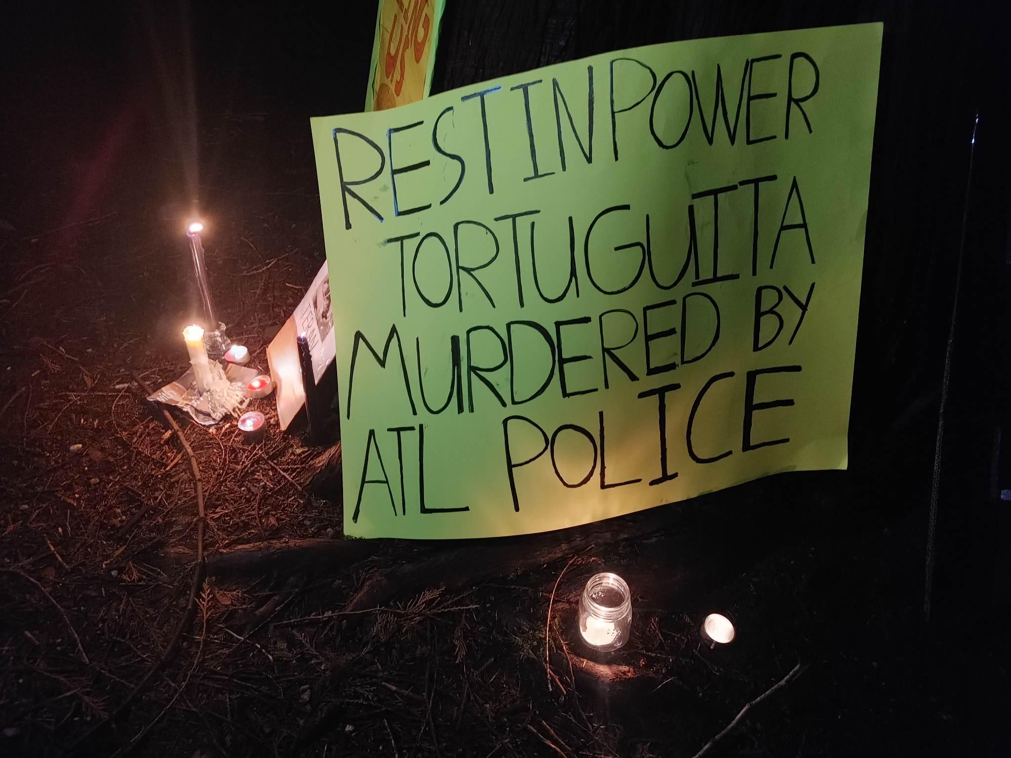 A green sign surrounded by candles reads "REST IN POWER TORTUGUITA MURDERED BY ATL POLICE" in black letters.
