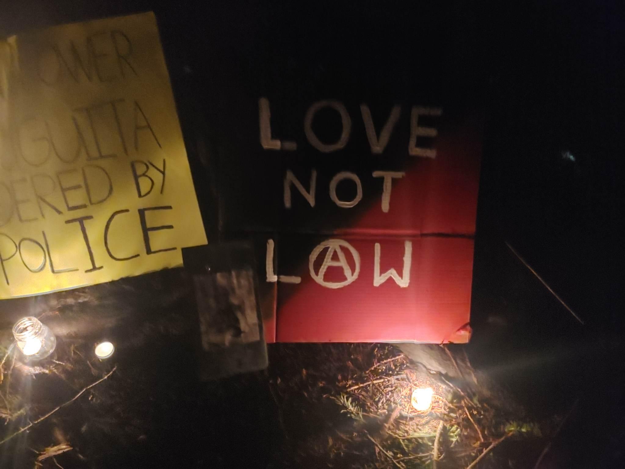 A sign with lit candles near it says "LOVE NOT LAW" in white font. The "A" in "Law" is an anarchist circle-A symbol. The background of the sign is a black and red anarcho-communist flag. 