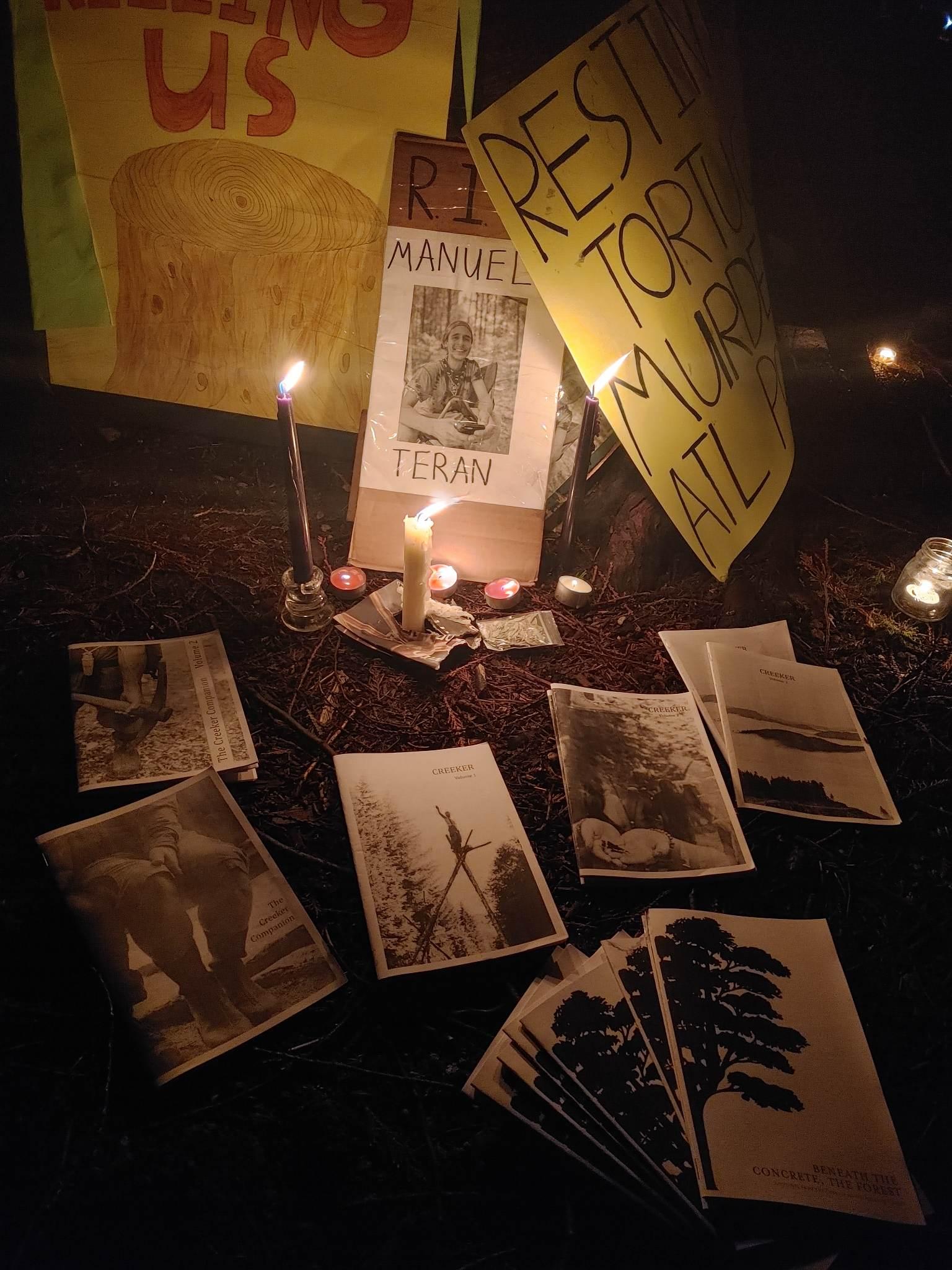 A picture of Tortuguita is lit by several vigil candles. Several zines surround the photo and candles. They include several "Creeker" zines, volumes 1-3. They also include both volume 1 and 2 of "The Creeker Companion." A stack of "Beneath the Concrete, The Forest" zines are also present.