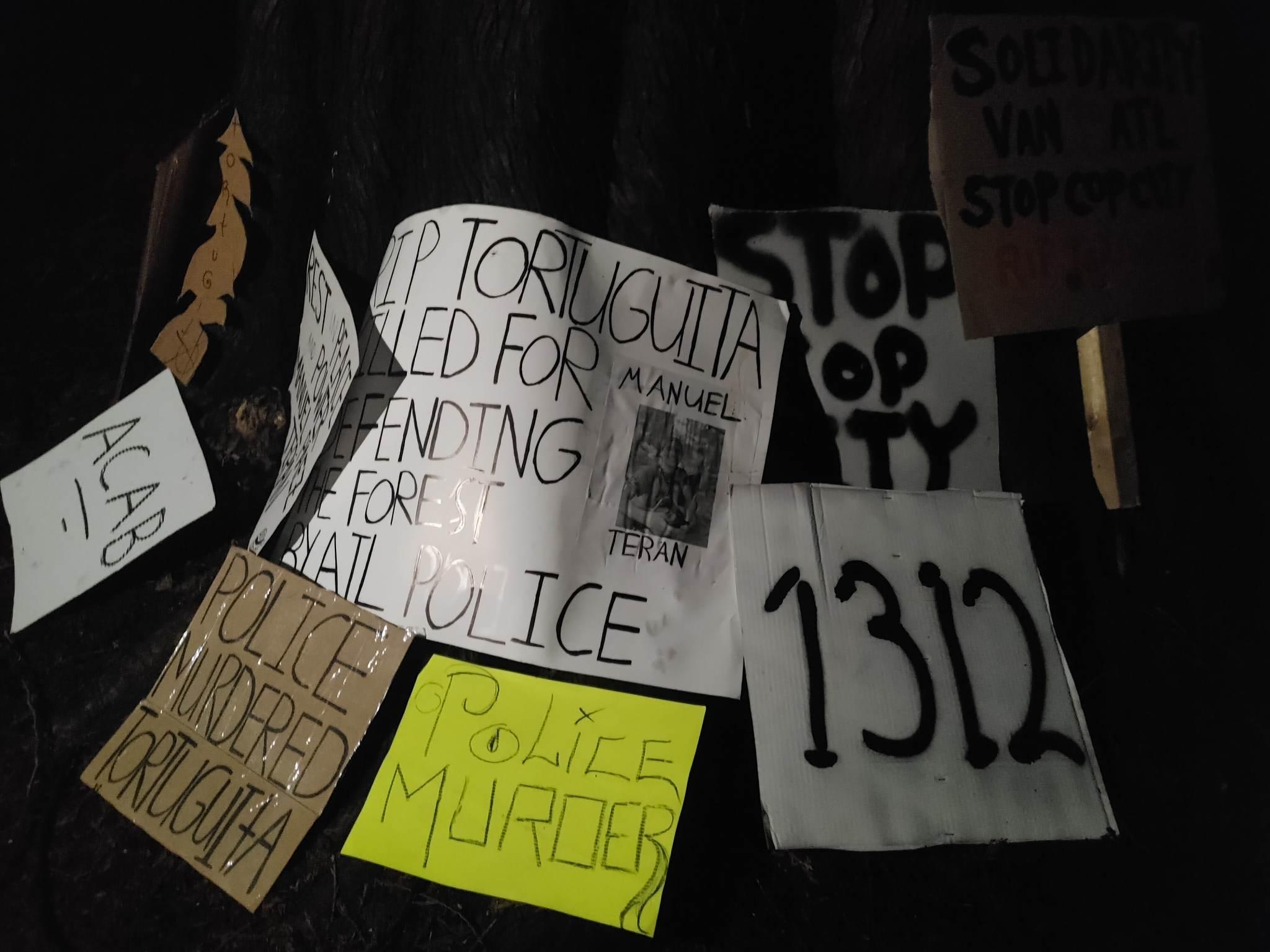 A stack of signs lay propped against a wall. They include "ACAB", "POLICE MURDERED TORTUGUITA", POLICE MURDER", "1312", "RIP TORTUGUITA KILLED FOR DEFENDING THE FOREST BY ATL POLICE" (this sign has a picture of Tortuguita and the name "Manuel Teran" on the right side), "STOP COP CITY", and a sign that says "SOLIDARITY VAN TO ATL STOP COP CITY RIP TORTUGUITA (the final sign has some text that is out of frame.)