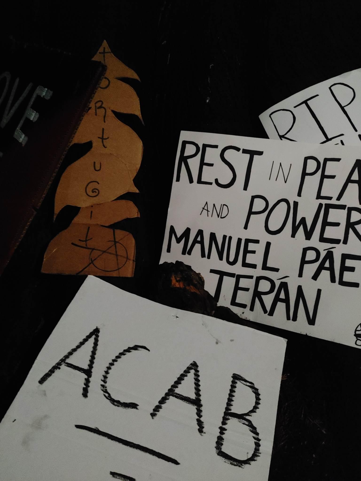 Several signs lay in a stack. One sign has a design of a tree and says "Tortuguita" with an anarchist circle-A symbol in the bottom right corner. The others say "ACAB", "REST IN PEACE AND POWER MANUEL PAEZ TERAN", and another is out of frame but the words "RIP" are visible.