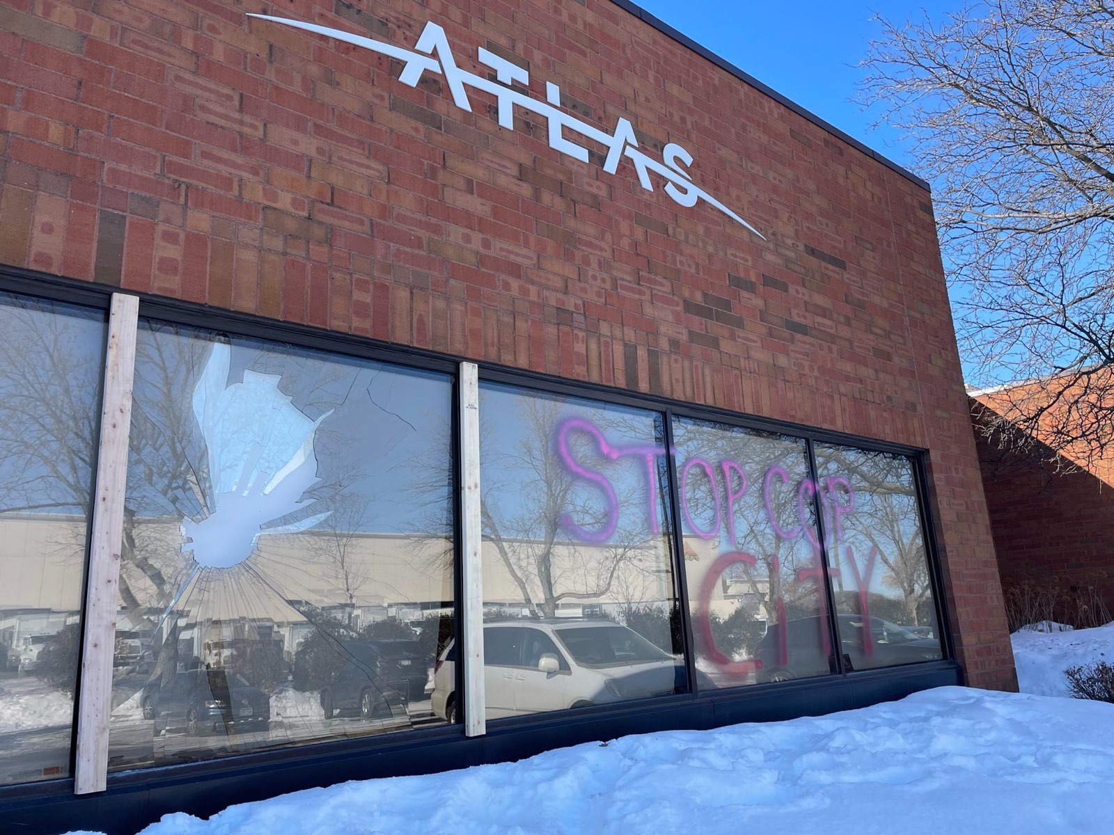 Under a brick wall with an Atlas logo, A glass window is broken. Three other glass windows are tagged "STOP COP CITY" in red spray paint.