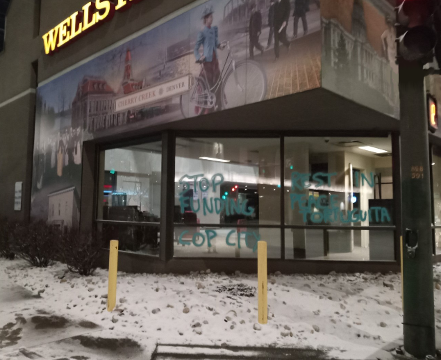 Two panes of glass on a wells fargo bank in a city with snow on the ground are spray painted. One says "STOP FUNDING COP CITY", another "REST IN PEACE TORTUGUITA." Both are tagged in green spray paint. There is a mural above the windows that depicts various scenes from the 18th through the 20th century that are difficult to discern.