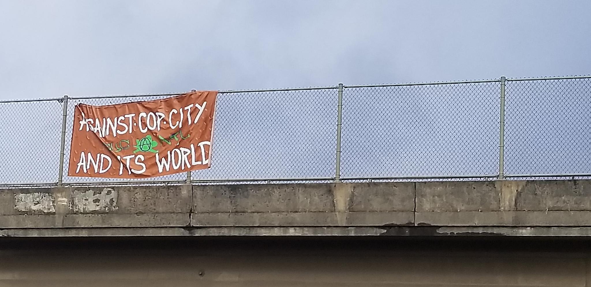 Onto a highway overpass, an orange banner with white text reads "AGAINST COP CITY AND ITS WORLD" with an anarchist circle-A in the center.