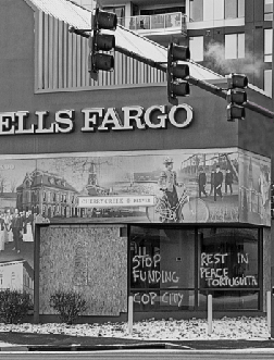 A grayscale photo of a bank. On the glass windows of a wells fargo bank underneath a mural, "STOP FUNDING COP CITY" is tagged on one pane of glass and "REST IN PEACE TORTUGUITA" is tagged on another.